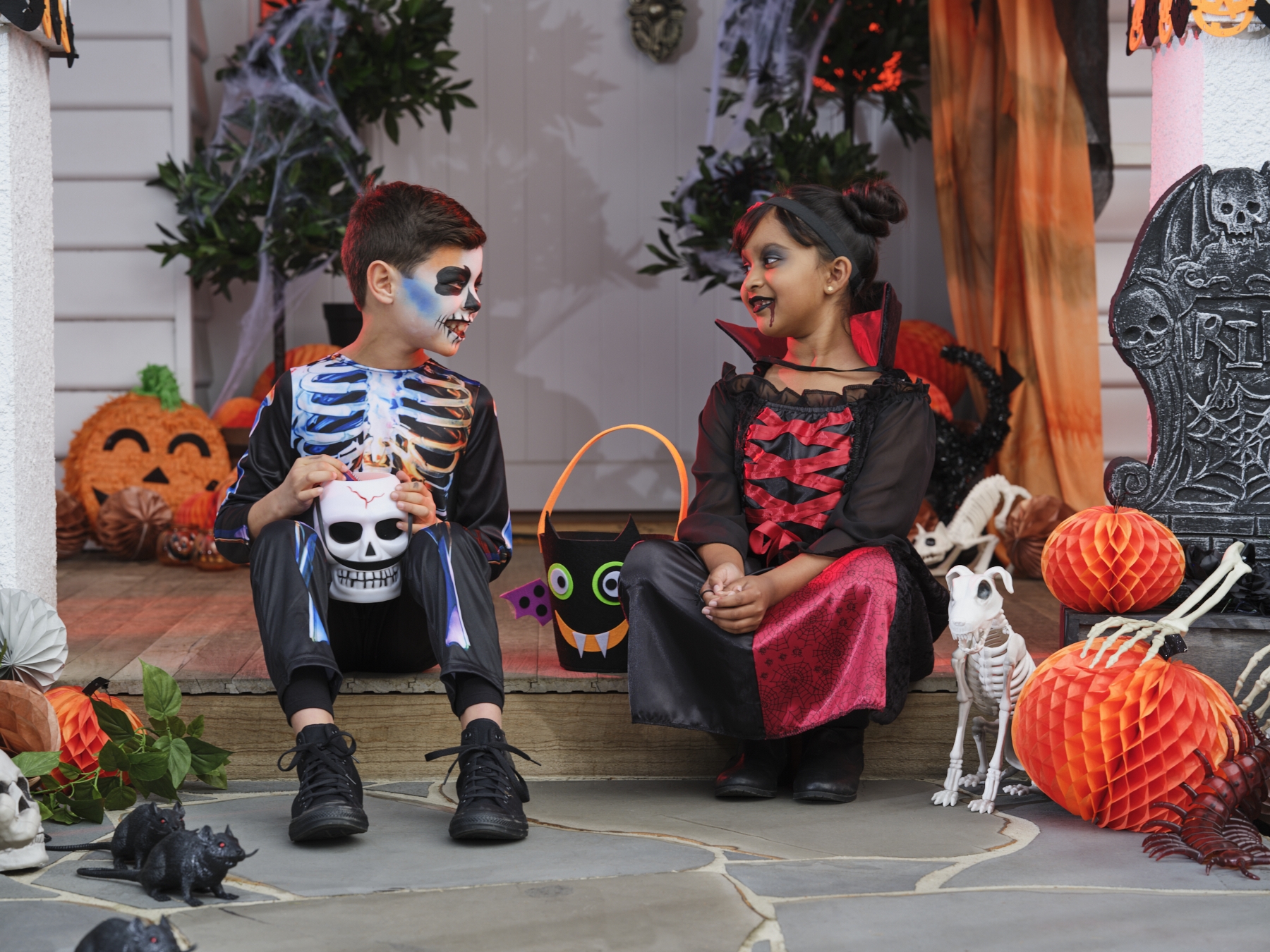 Two kids in Halloween costumes sitting on a front porch step decorated with pumpkins, skeletons and cobwebs